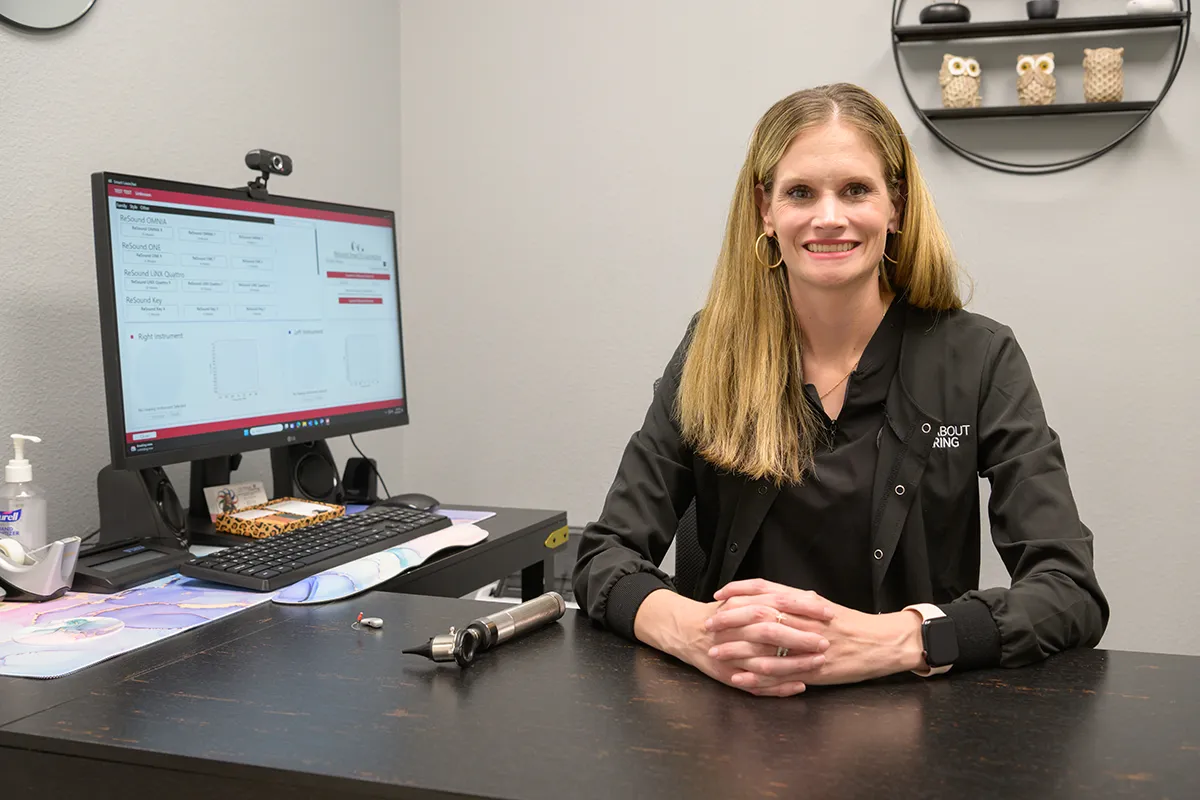 Dr. Guempel smiling at her desk in an All About Hearing jacket, next to a hearing aid fitting screen and an otoscope